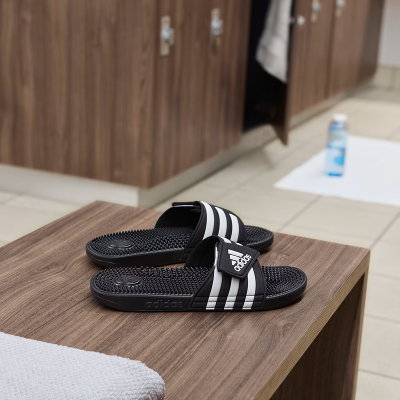 Adidas Adissage Slide sandals in black with white stripes and massaging footbed, side view placed on wooden bench.