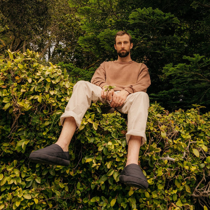 "SeaVees Apres Puff Slip On shoes in black with quilted design and cushioned construction, worn outdoors on a lush greenery backdrop"