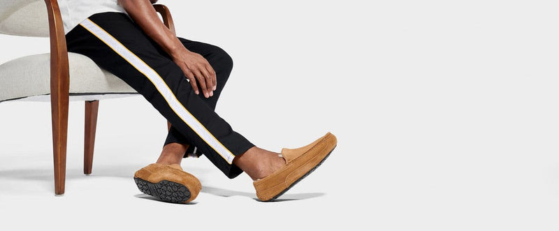 UGG Ascot Suede slippers in chestnut suede with cushioned sole and moccasin-style stitching, worn in lifestyle sitting context.