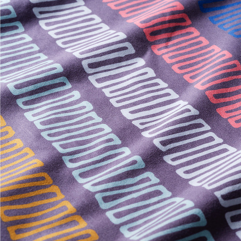 Nomadix Bandana Towel featuring geometric patterns in blue, orange, and red on a purple background, close-up view.