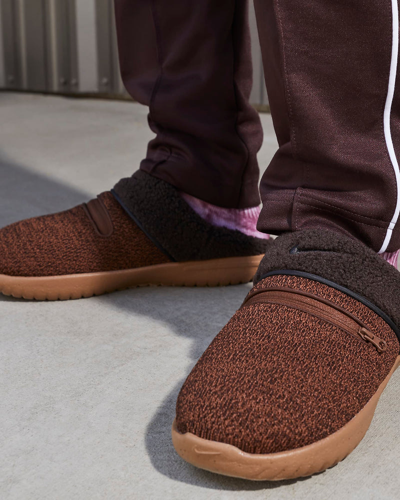 Nike Burrow SE slippers in brown with textured knit upper, zippered top detail, fleece lining, and rubber outsole, lifestyle context