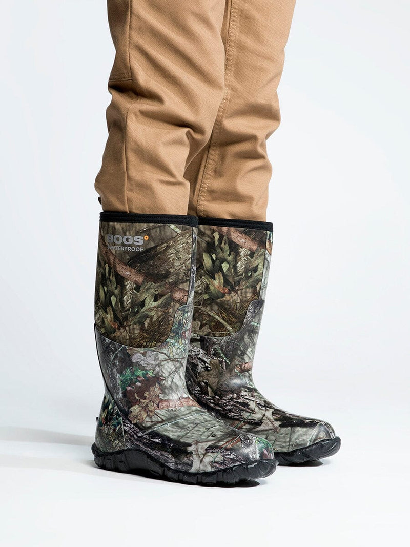 Bogs Classic High Mossy Oak waterproof boots with camouflage print and reinforced rubber soles, side view worn by person.
