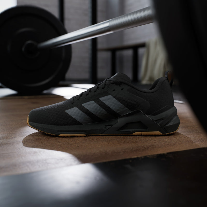 Adidas Dropset Control Trainer shoes in black with textured upper, gray stripes, and gum sole, side view placed in a gym setting.