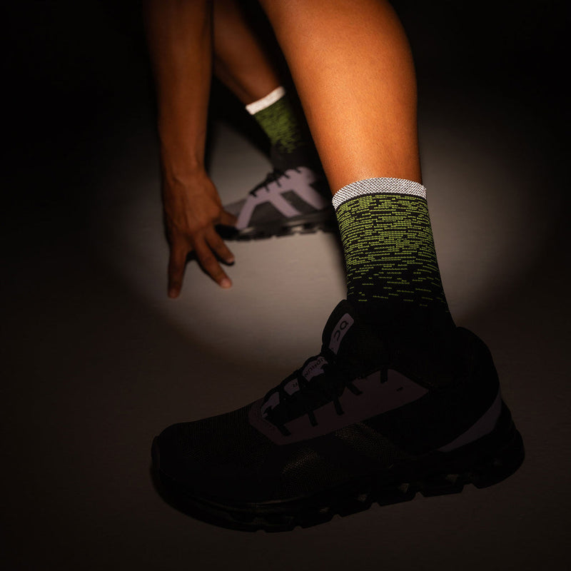 Feetures Elite Light Cushion Reflective Mini Crew socks in black and green with reflective cuff, worn by athlete in stretching pose.