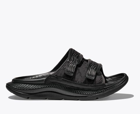 Side view of the HOKA ORA Luxe recovery slide in black. Features an adjustable Ariaprene foam and mesh strap, a cushioned sugarcane EVA midsole, and a lightweight design for post-run comfort.