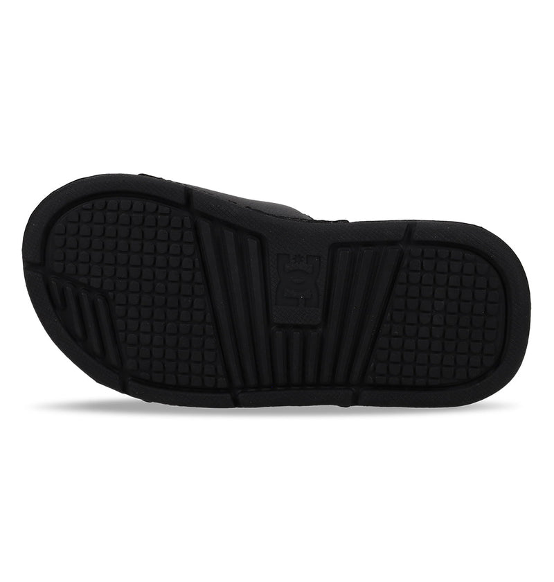 The image shows the black rubber sole of DC Shoes Kids' Bolsa Slides, featuring square-patterned treads and grooves for grip, plus a raised logo in the center—ideal for comfy kids slide sandals.