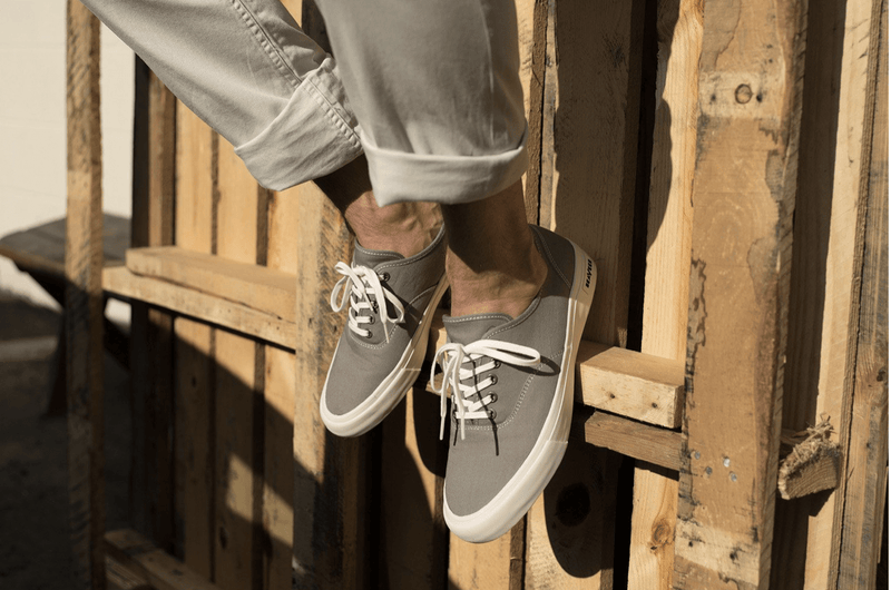 SeaVees Legend Sneaker Standard in gray with white laces and rubber sole, casual lifestyle worn sitting on wooden pallets.