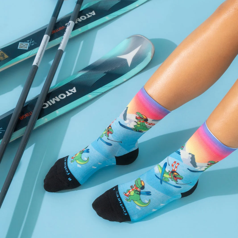 Feetures Limited Edition "Ski-Rex" Elite Light Cushion Mini Crew socks in blue with colorful dinosaur skiing graphics, worn lifestyle view.