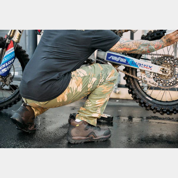 DC WorkCrew Navigator Work Hi Top Comp Toe with Met Guard work boots in brown with reinforced toe and rugged outsole, worn by a man crouching near a motorcycle.