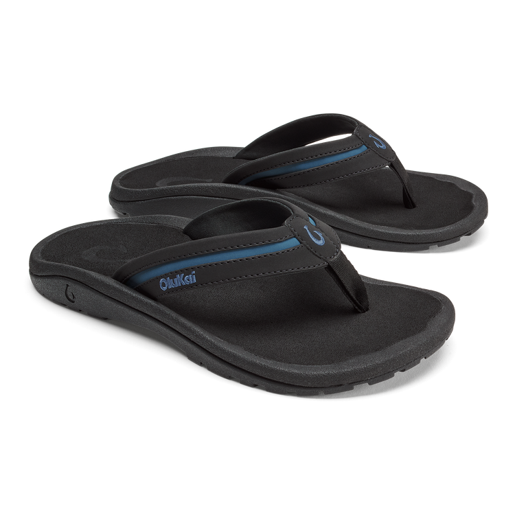 OluKai ʻOhana Pālua sandals featuring a black footbed, dark straps with a blue accent, and a durable rubber outsole, angled side view.