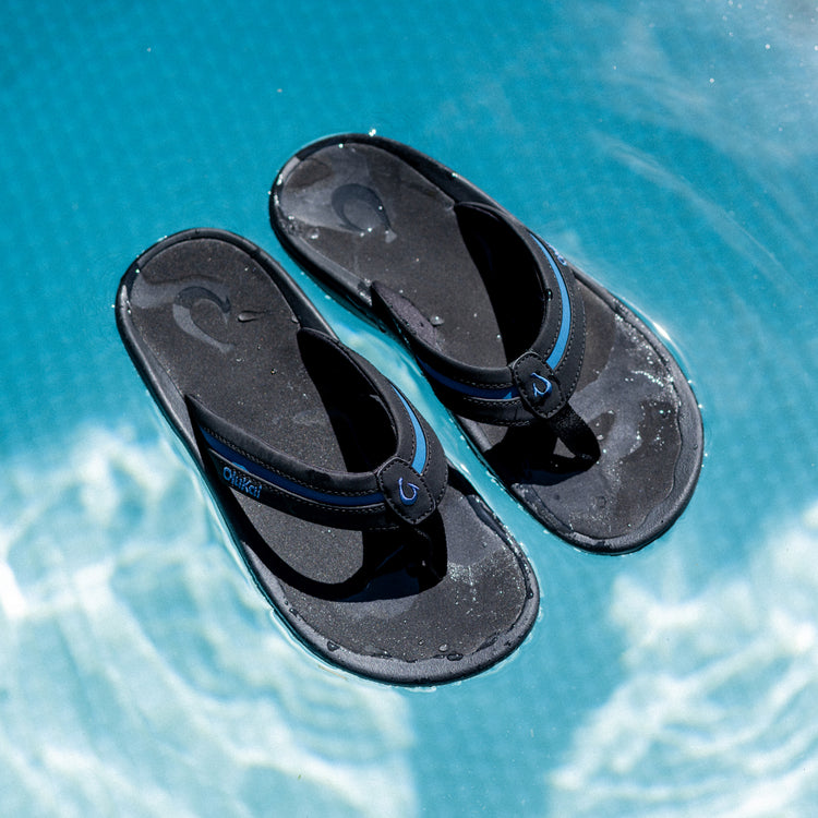 OluKai ʻOhana Pālua sandals in black with blue accents, water-resistant design and contoured footbed, top view on a pool surface.