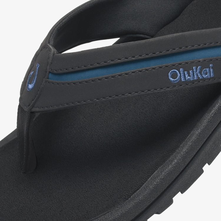OluKai ʻOhana Pālua sandals in black with blue embroidered logo and textured footbed, close-up angled view.