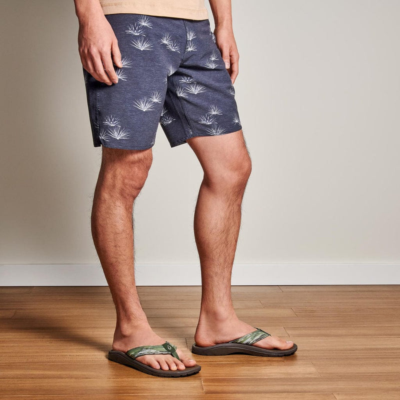 OluKai Ohana Pa'i sandals in green with tropical print straps and cushioned brown footbed, worn by a person indoors.