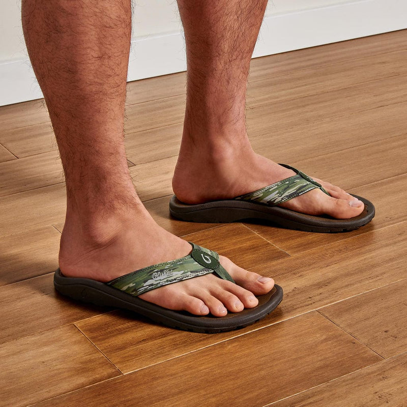 "OluKai Ohana Pa'i sandals with green patterned straps and brown cushioned soles worn by a person on wooden flooring"