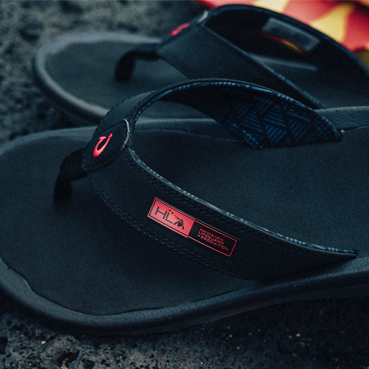 OluKai ‘Ohana sandals in black featuring red logo detail, textured straps, and cushioned footbed, angled close-up view.