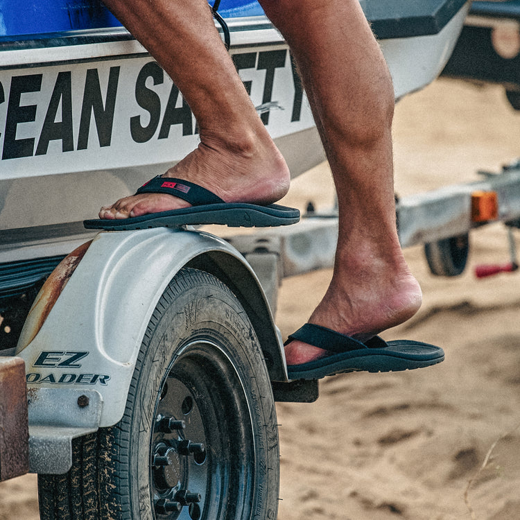 OluKai 'Ohana sandals in black with water-resistant straps and contoured footbed, lifestyle view worn near a boat trailer.