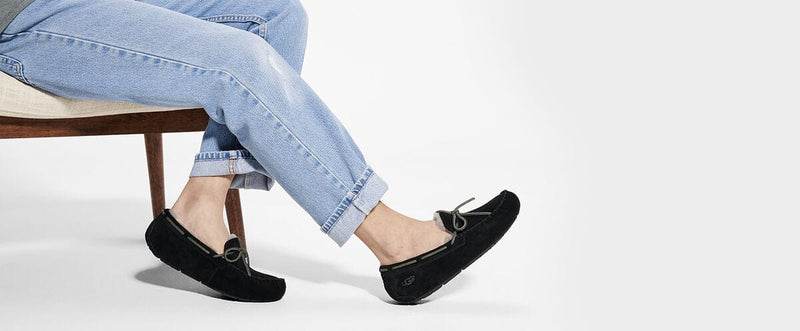 UGG Olsen moccasin slippers in black suede with bow detail and shearling lining, worn by seated individual.