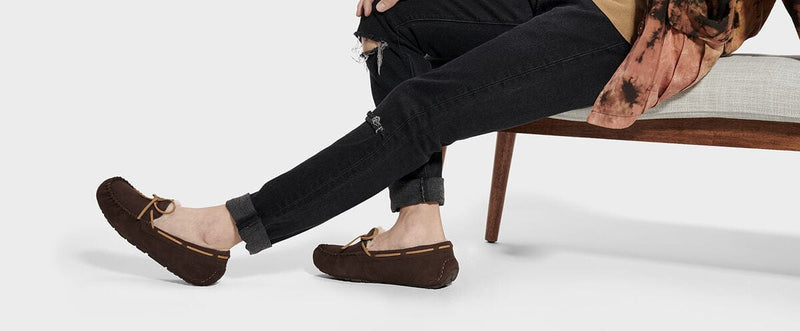 "UGG Olsen slippers in brown suede with leather laces and moccasin stitching, worn by person sitting on a chair"