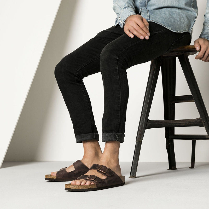 Birkenstock Arizona Oiled Leather sandals in dark brown with double adjustable straps and cork footbed, worn by seated individual.