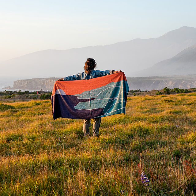 Nomadix Original Towel Big Sur featuring a colorful landscape design in orange, blue, and green, worn by a person outdoors.