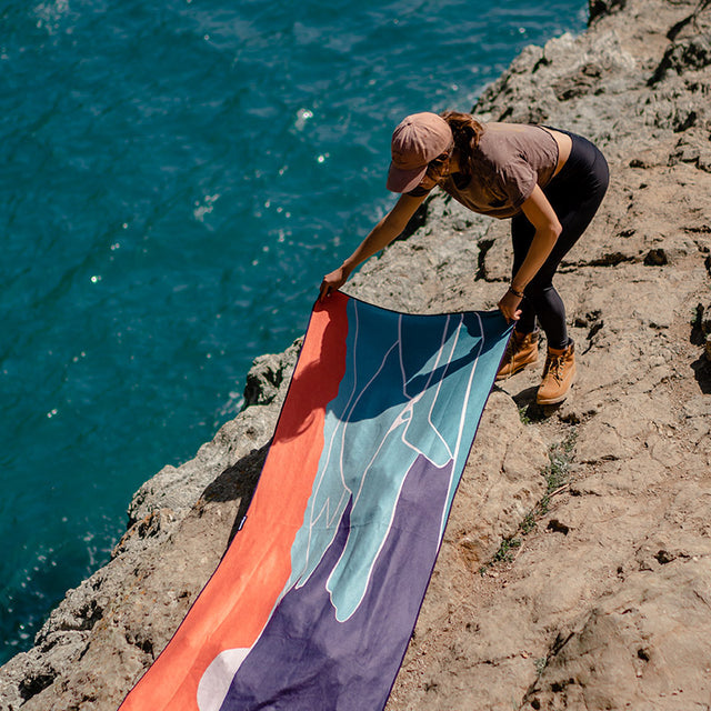 Nomadix Original Towel Big Sur, quick-drying towel with colorful abstract design featuring blue, orange, and red tones, lifestyle setting near ocean.