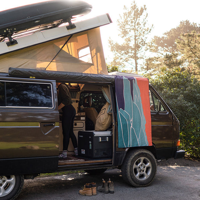 Nomadix Original Towel Big Sur, a multi-colored eco-friendly towel featuring a vibrant abstract design, draped over a van door outdoors.