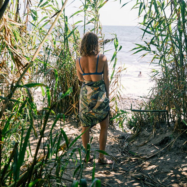 Nomadix Original Towel with tropical leaf print in green and beige tones, worn as a wrap by a person walking toward a beach.