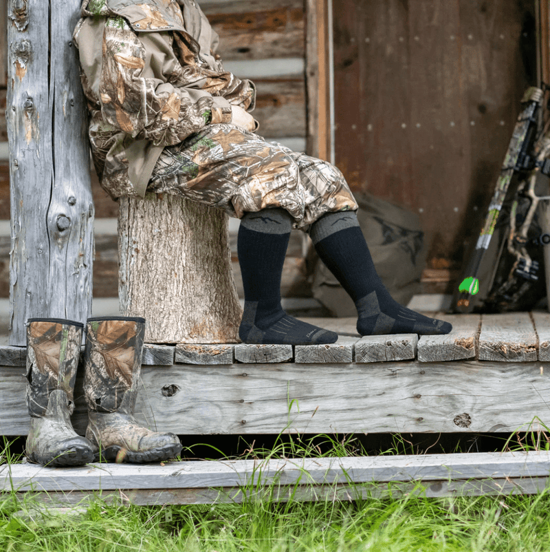 Darn Tough Over-the-Calf Heavyweight Hunting Sock in black with reinforced cushioning, worn outdoors in hunting setting.