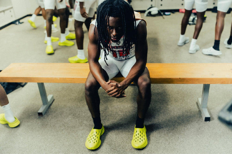 Kane Revive OB recovery shoes in bright yellow with perforated design and slip-on style, worn by an athlete in a locker room.