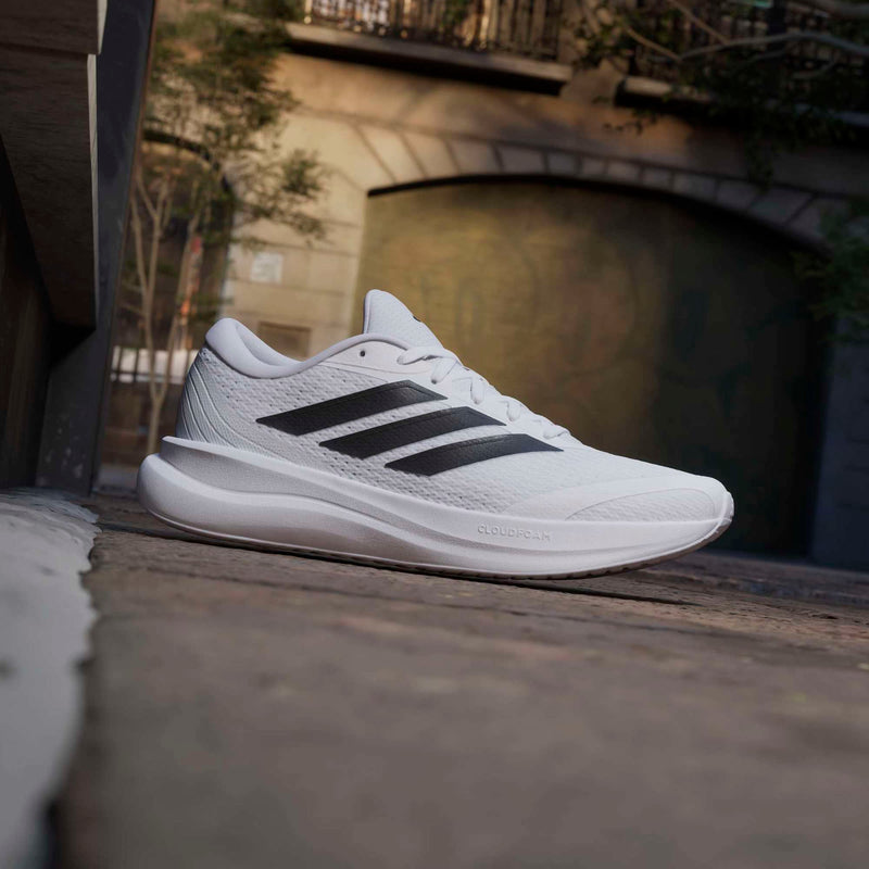 Adidas Runfalcon 6 running shoe in white with black stripes and Cloudfoam cushioning, angled side view on urban street.