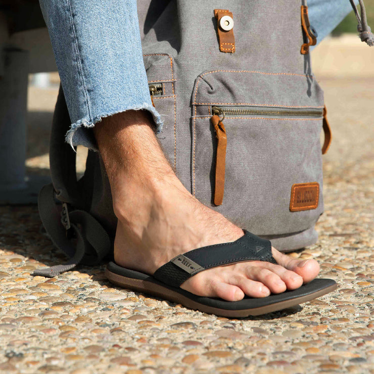 Reef Santa Anna sandals in black with ergonomic straps and cushioned footbed, worn outdoors on a pebble pathway.