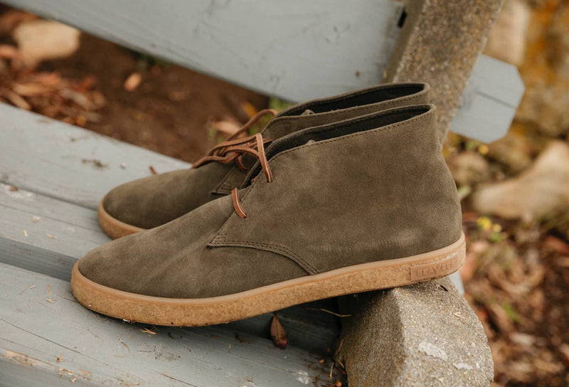 SeaVees Sun-Tans Chukka shoes in olive green suede with tan rubber soles and brown laces, side view on outdoor bench.
