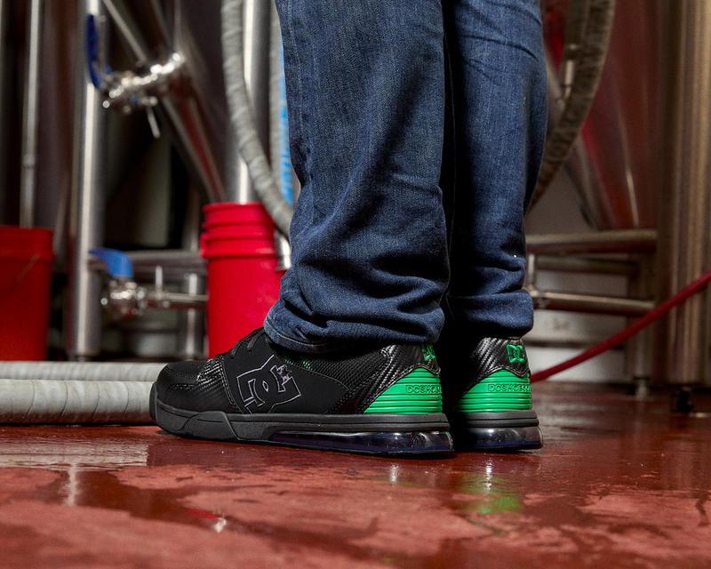 DC WorkCrew Versatile Work Comp Toe work shoes in black with green heel accents and breathable mesh material, worn in an industrial setting.