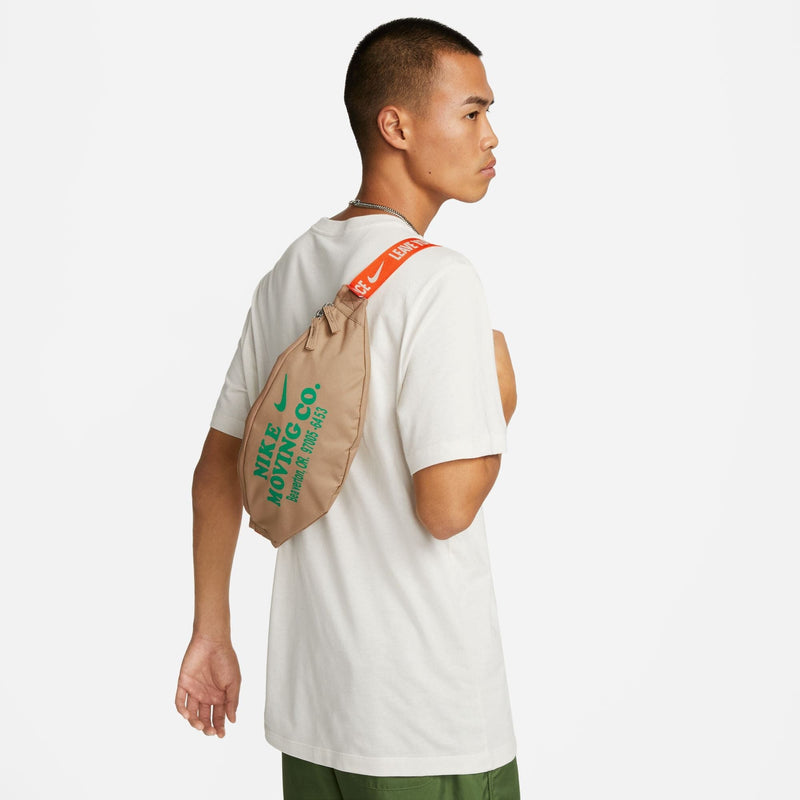 Nike Waist Pack in beige with green "Nike Moving Co." graphic and bright orange adjustable strap, worn over the shoulder.